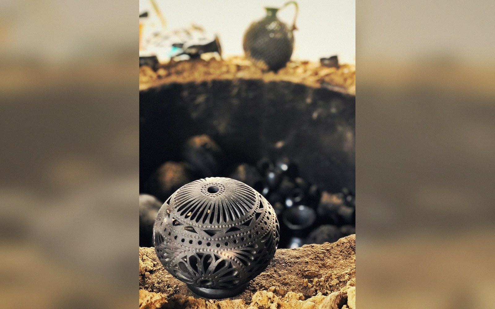 Intricately carved black clay sphere displayed beside a pit kiln with a jug