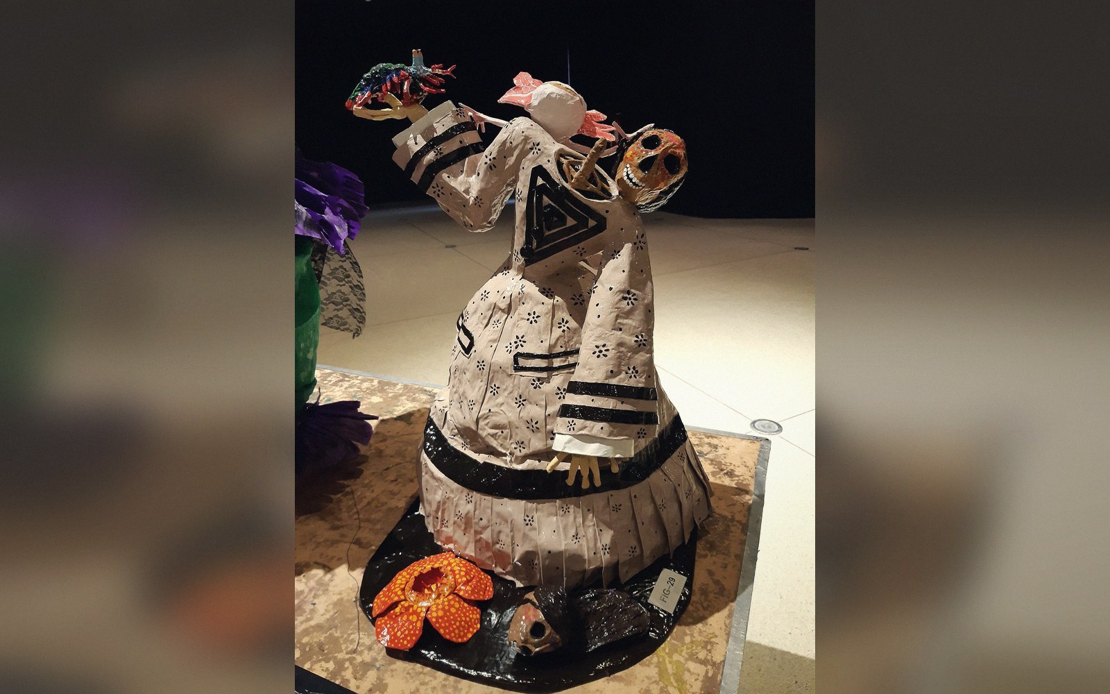 Sculptural papier-mâché figure with painted patterns, holding animal forms, displayed on a pedestal