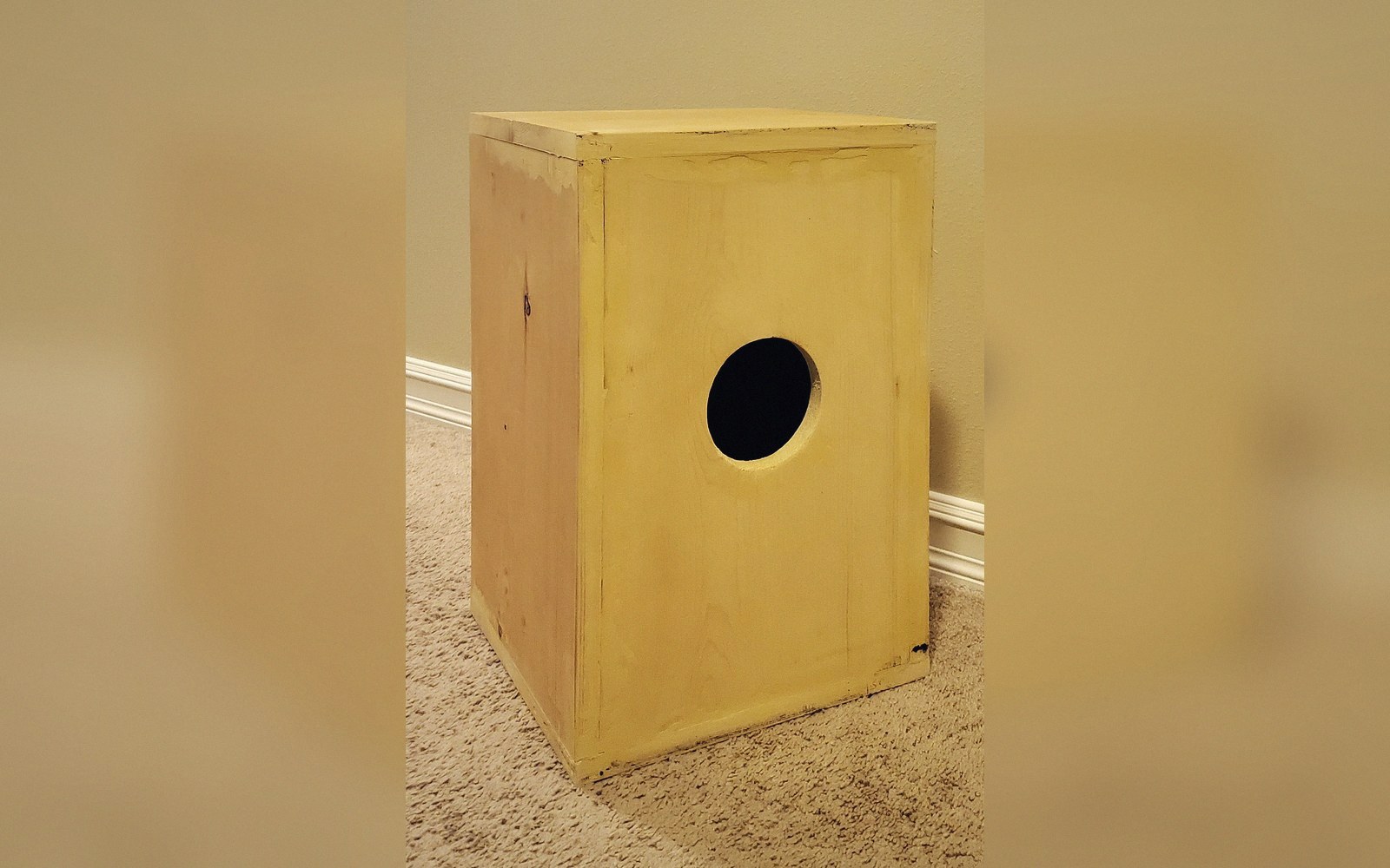 A wooden cajón drum box with a round sound hole, standing on carpeted floor