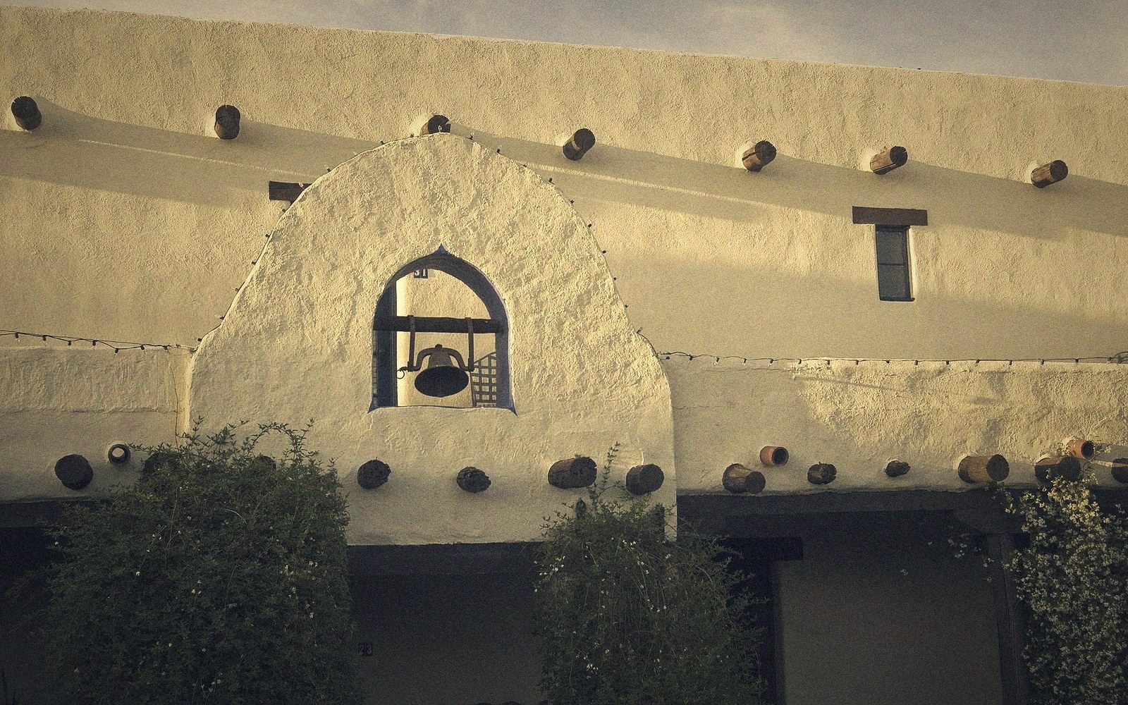 Adobe-style facade with a bell mounted in an arched niche above a shaded doorway
