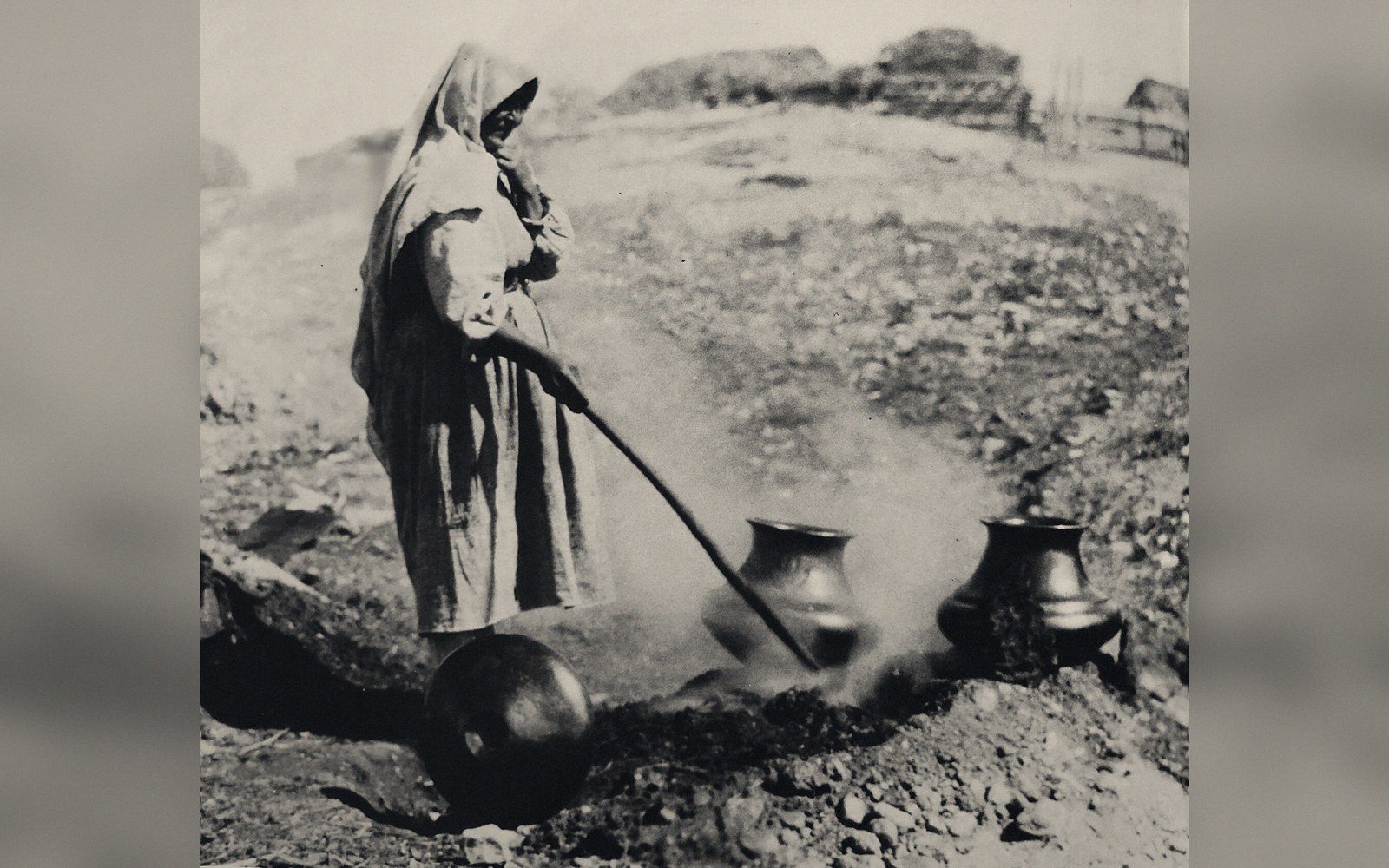 A hooded figure tends smoking clay pots buried in an open-air firing pit