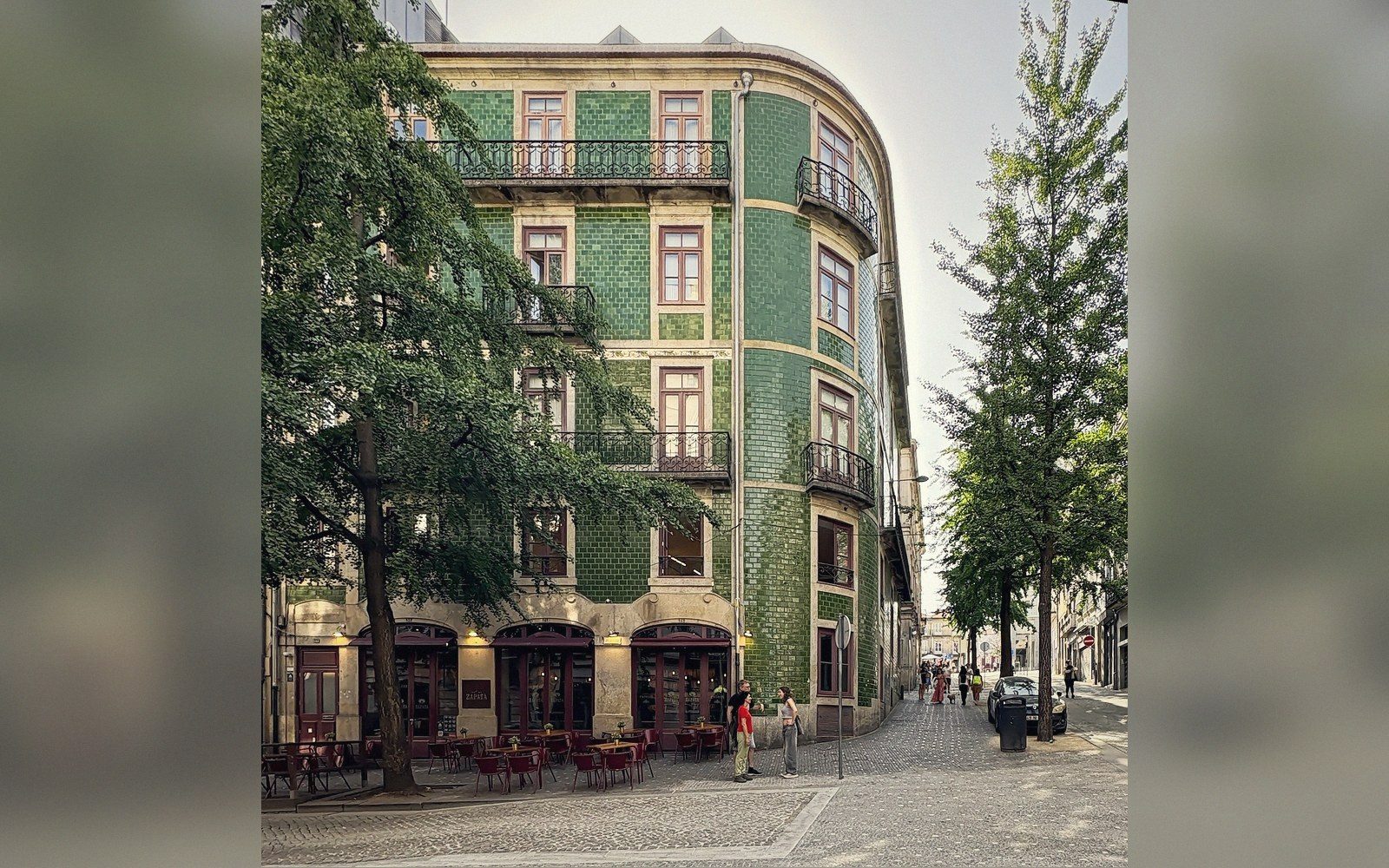 Rounded corner building with green glazed tile facade and a ground-floor café on a cobblestone street
